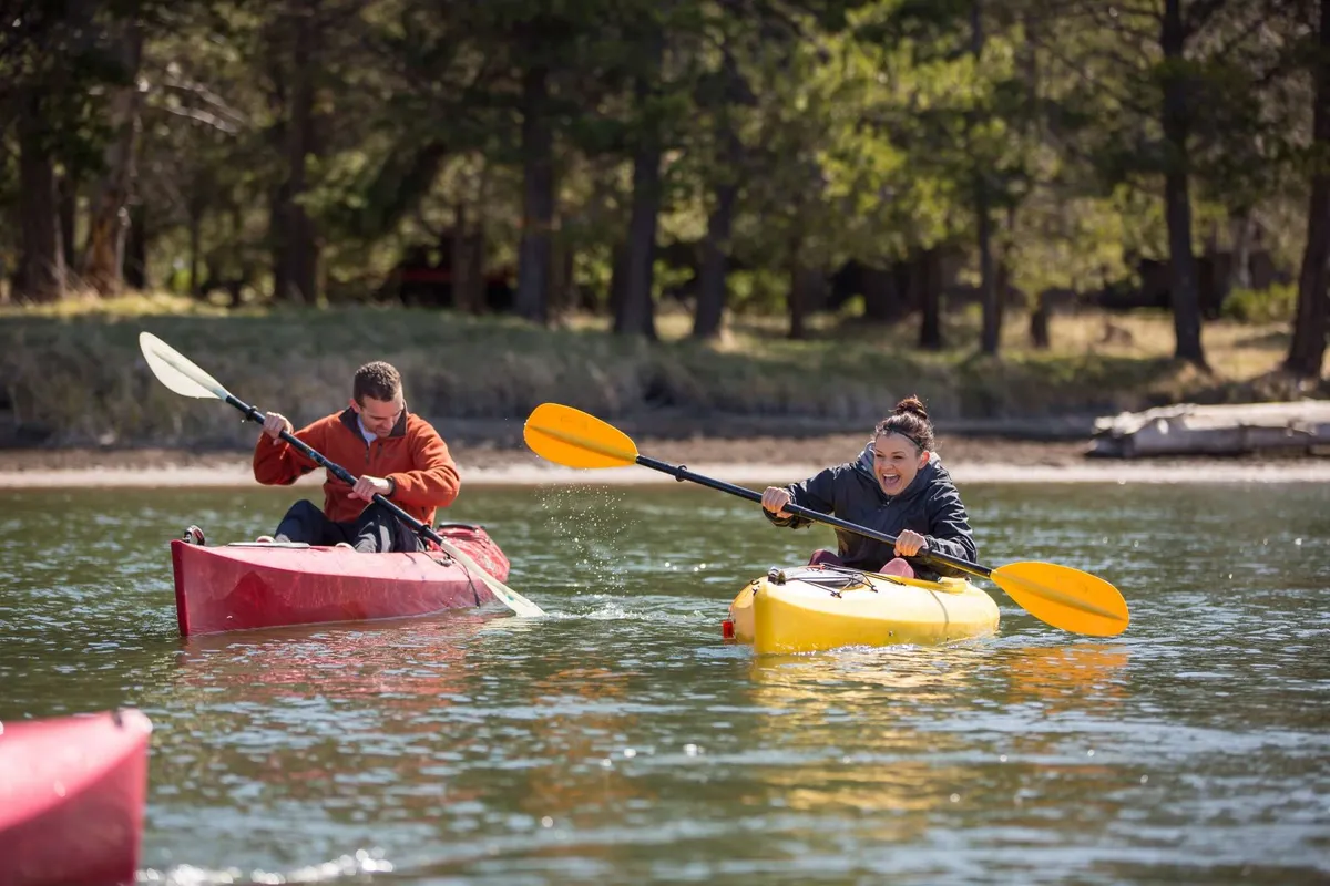 Two kayakers glide across a calm, tree-lined river, their brightly colored kayaks adding pops of color to the tranquil natural setting. The vibe is playful and energetic, highlighting active outdoor recreation.