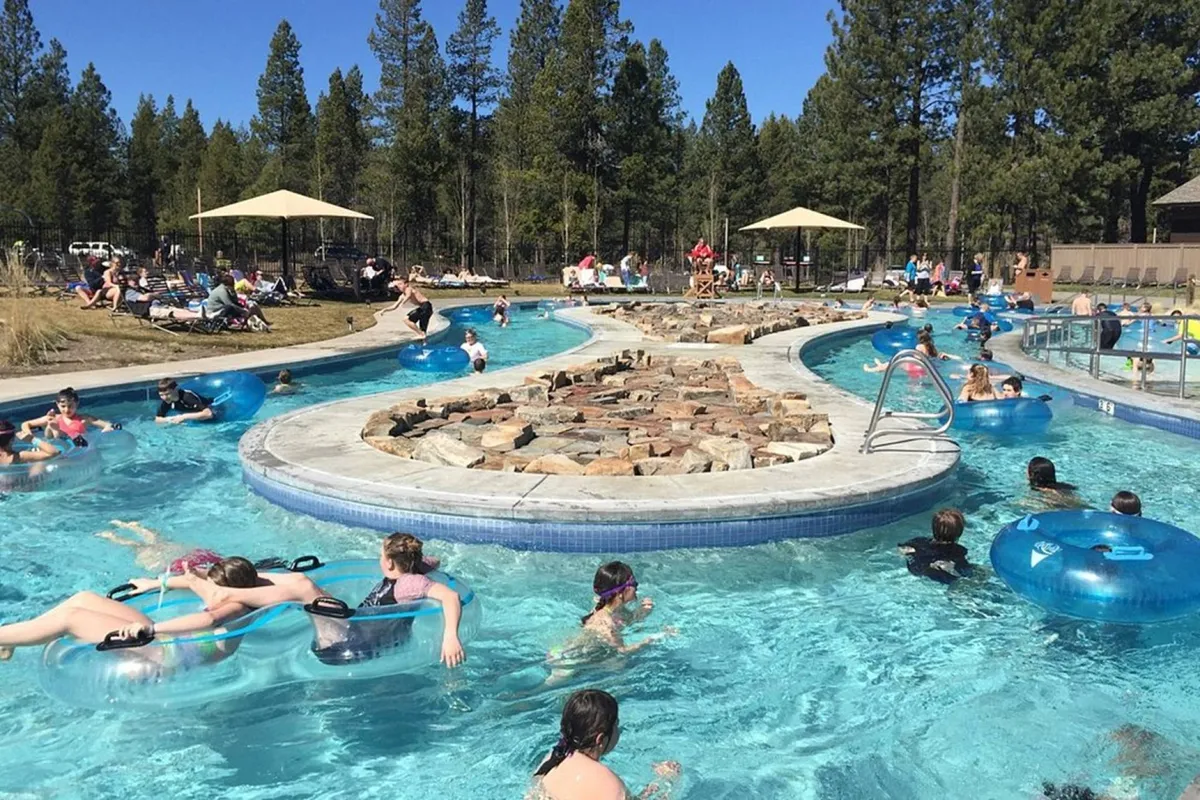 A lively, sunlit swimming pool scene featuring people floating in the water, natural rock formations, and colorful sun umbrellas. The atmosphere is joyful and full of family energy, evoking the carefree fun of a community aquatic center.