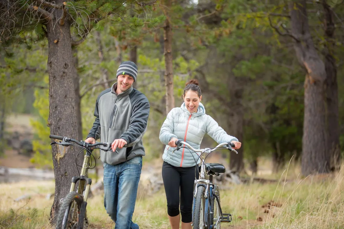 A couple walks their bicycles beneath tall trees across a lush, open grassy area. The sun filters through the leaves, and the setting suggests immediate access to scenic bike trails and the ease of exploring Sunriver’s natural beauty.