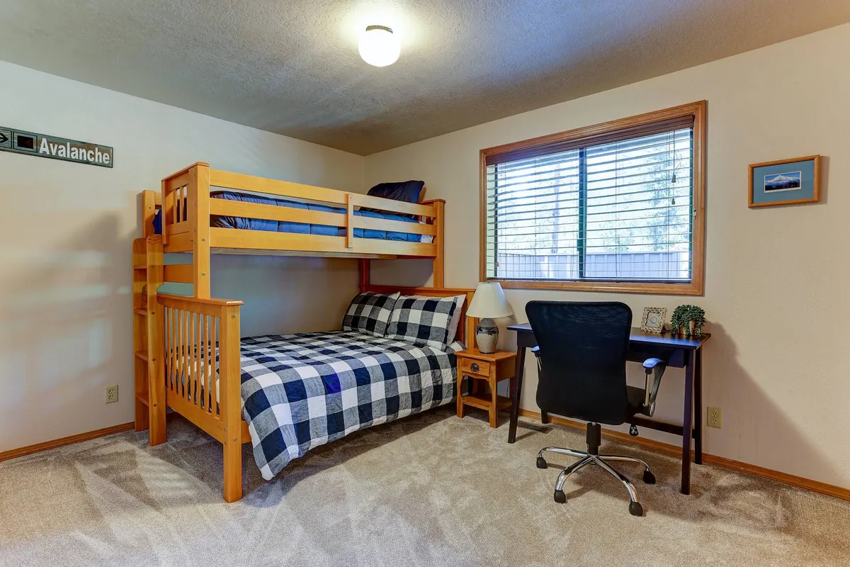 A cheerful bunk room with a sturdy wooden bunk bed outfitted in plaid bedding, a desk and chair set up by the window, and ample natural light. The space feels inviting and practical, suitable for kids or friends.