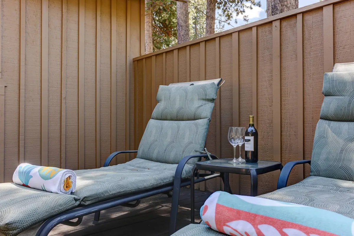 Two lounge chairs are positioned outdoors on a deck or patio, with a small side table holding wine and glasses nearby. Colorful towels add a vibrant touch, and the setting suggests relaxation under the sun or stars.