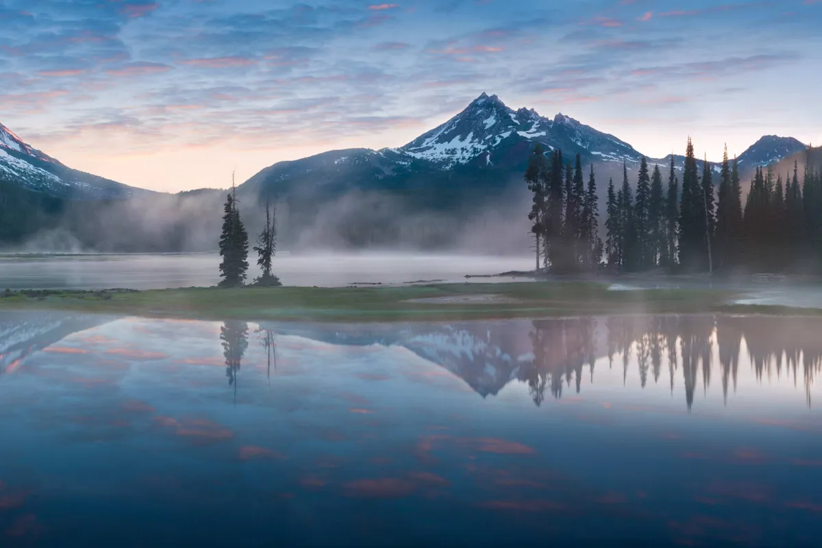 A sweeping view of a tranquil lake surrounded by lush pine trees and distant mountains under a wide, open sky. The scene captures the vastness and natural beauty of Central Oregon, inviting activities like hiking, fishing, and wildlife watching.