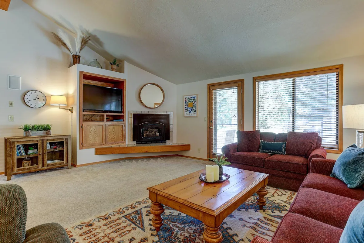 A cozy living area with a plush sofa arranged around a gas fireplace set in a rustic rock hearth. Large windows let in ample natural light, and a stylish coffee table anchors the seating area—perfect for relaxing, hosting game nights, or gathering for conversations.