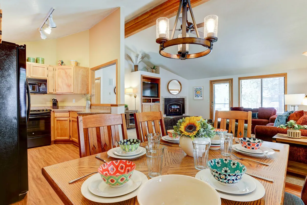 An open-concept space featuring a dining table set with colorful bowls and a chandelier overhead. The view extends into the kitchen area and living space, showcasing how the dining, kitchen, and relaxation zones seamlessly flow together.