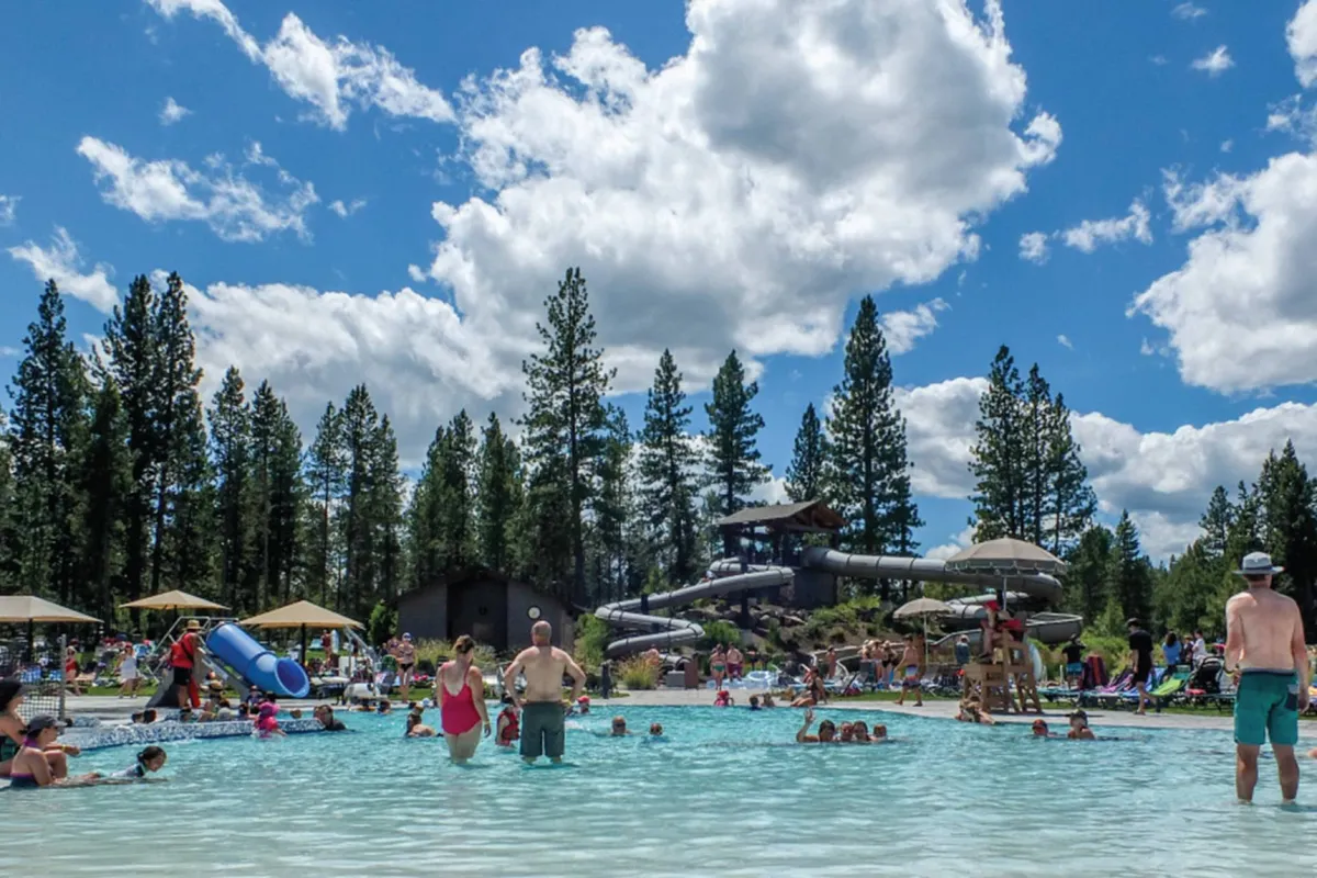 An expansive outdoor pool area with a dynamic water slide, sun loungers, swimmers of all ages, and lush trees in the background. The mood is festive and carefree, with people laughing, playing, and relaxing by the water.
