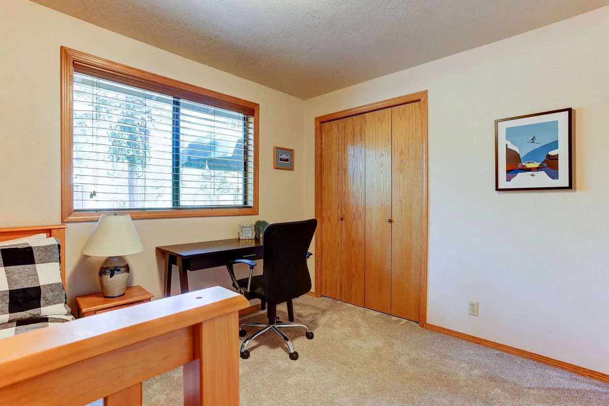 A bedroom with a comfortable bed draped in a checkered blanket, a window with blinds letting in natural light, and a dedicated workspace featuring a desk and chair. Framed artwork adds personality to the space.