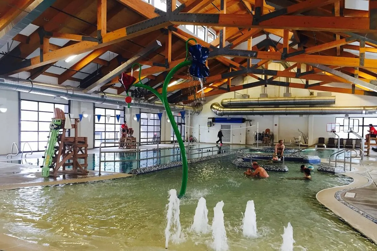 An indoor aquatic center featuring a sparkling swimming pool, twisting water slides, playful fountain features, and sturdy wooden beams overhead. Families and children are seen swimming, playing, and relaxing, creating a lively, joyful atmosphere.
