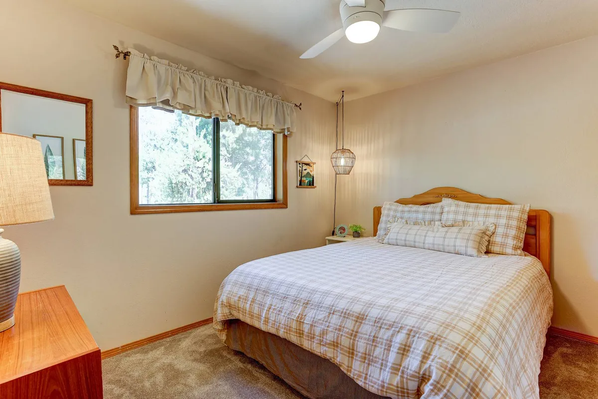 A restful bedroom with a queen-sized bed dressed in a plaid comforter, a large window streaming in gentle natural light, and a wooden nightstand with a lamp. The space is thoughtfully arranged for comfort, with a hanging light fixture adding a touch of warmth.