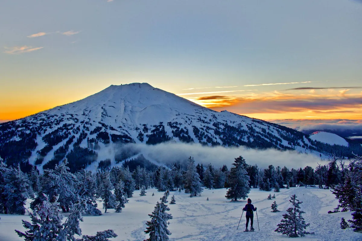 A snow-capped mountain dominates the background, with the silhouette of a skier gliding in the foreground and pine trees below. The scene captures the grandeur and adventure of winter sports in the Sunriver region.
